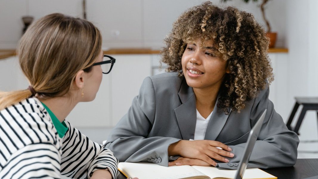 Black professional feeling overwhelmed at her desk while colleagues work nearby.
